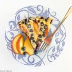 A slice of Gluten-Free Blueberry Peach Clafoutis on a blue patterned plate with fresh peach slices