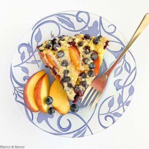 A slice of Gluten-Free Blueberry Peach Clafoutis on a blue patterned plate with fresh peach slices