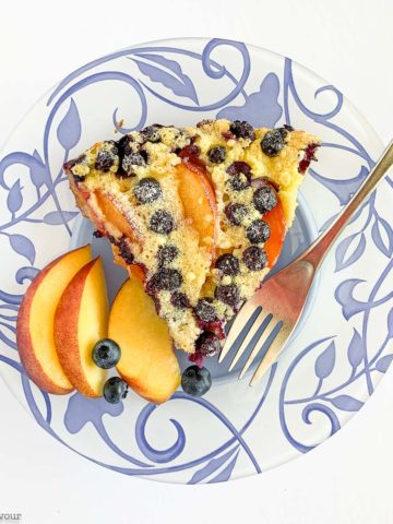 A slice of Gluten-Free Blueberry Peach Clafoutis on a blue patterned plate with fresh peach slices