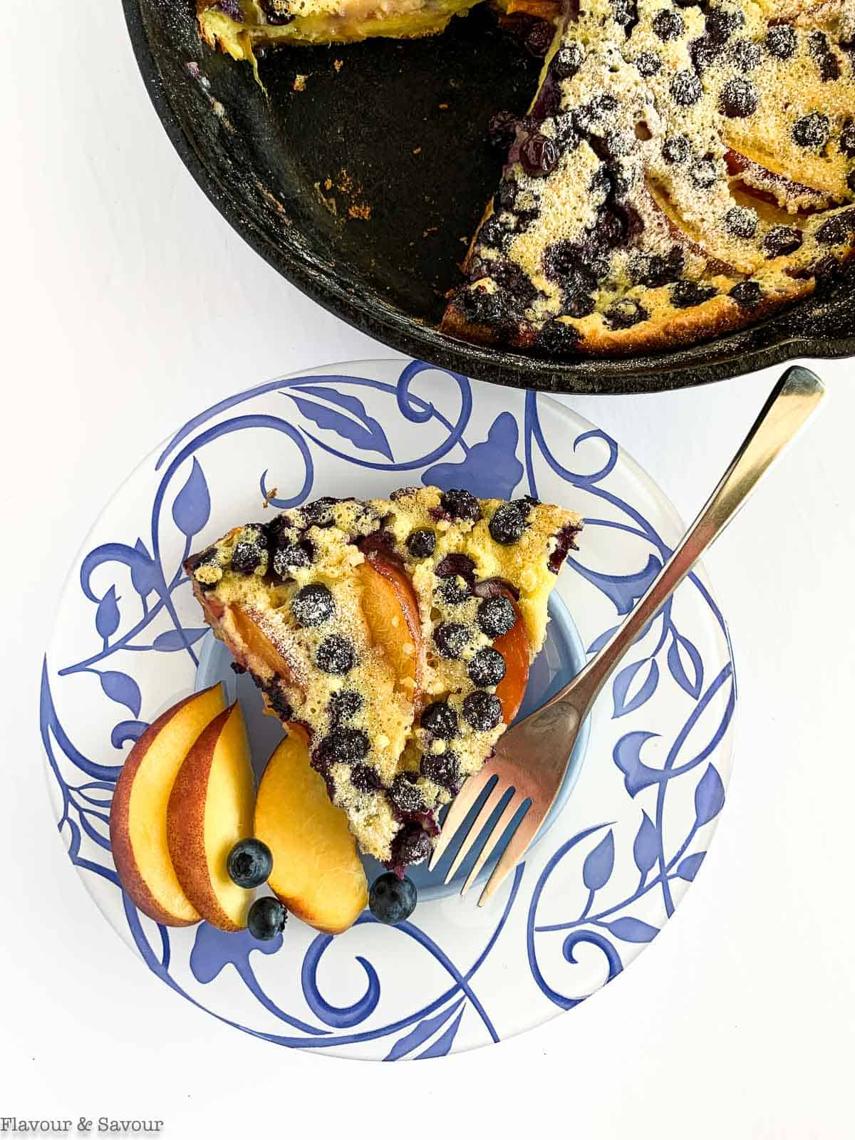 Overhead view of Gluten-Free Blueberry Peach Clafoutis on a plate beside a cast iron skillet