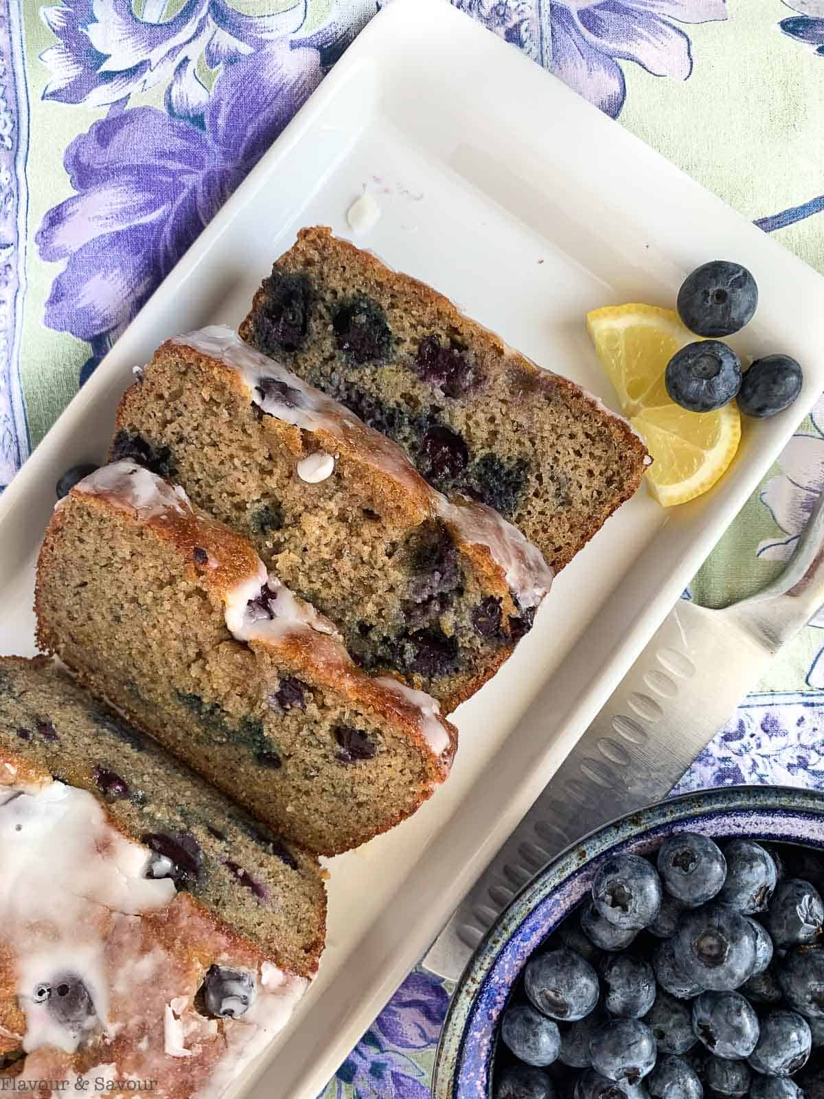 Slices of Blueberry Banana Bread on a white plate with a bowl of blueberries nearby.