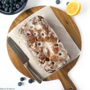 A loaf of glazed blueberry banana bread on a round wooden board with a bowl of blueberries.