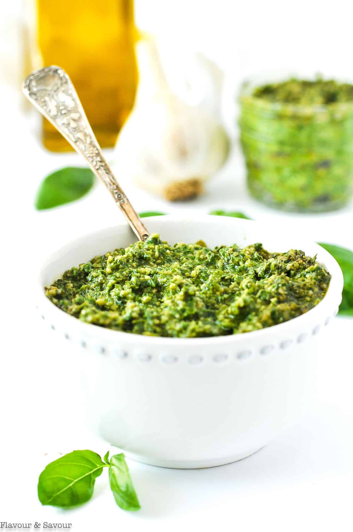 A small white bowl filled with Basil Cashew Pesto with garlic and olive oil in the background