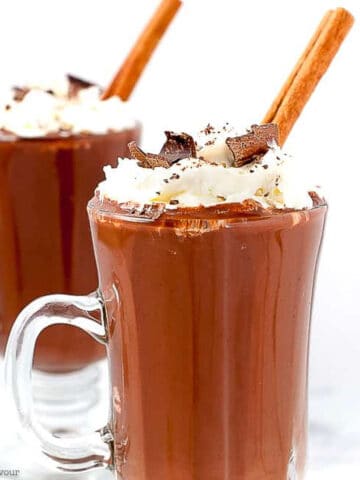 Two mugs of Mexican Hot Chocolate with whipped cream and cinnamon sticks.