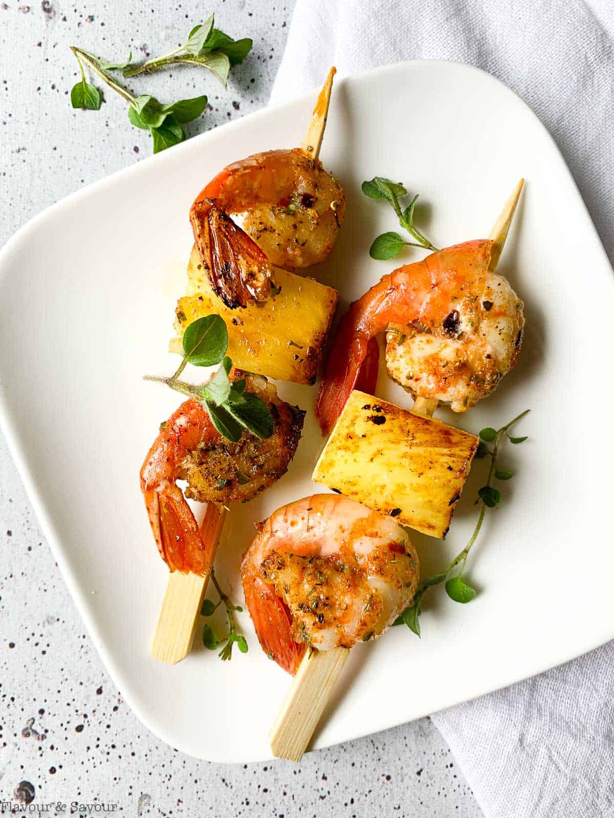Close up view of two skewers of prawns and pineapple with fresh oregano leaves.
