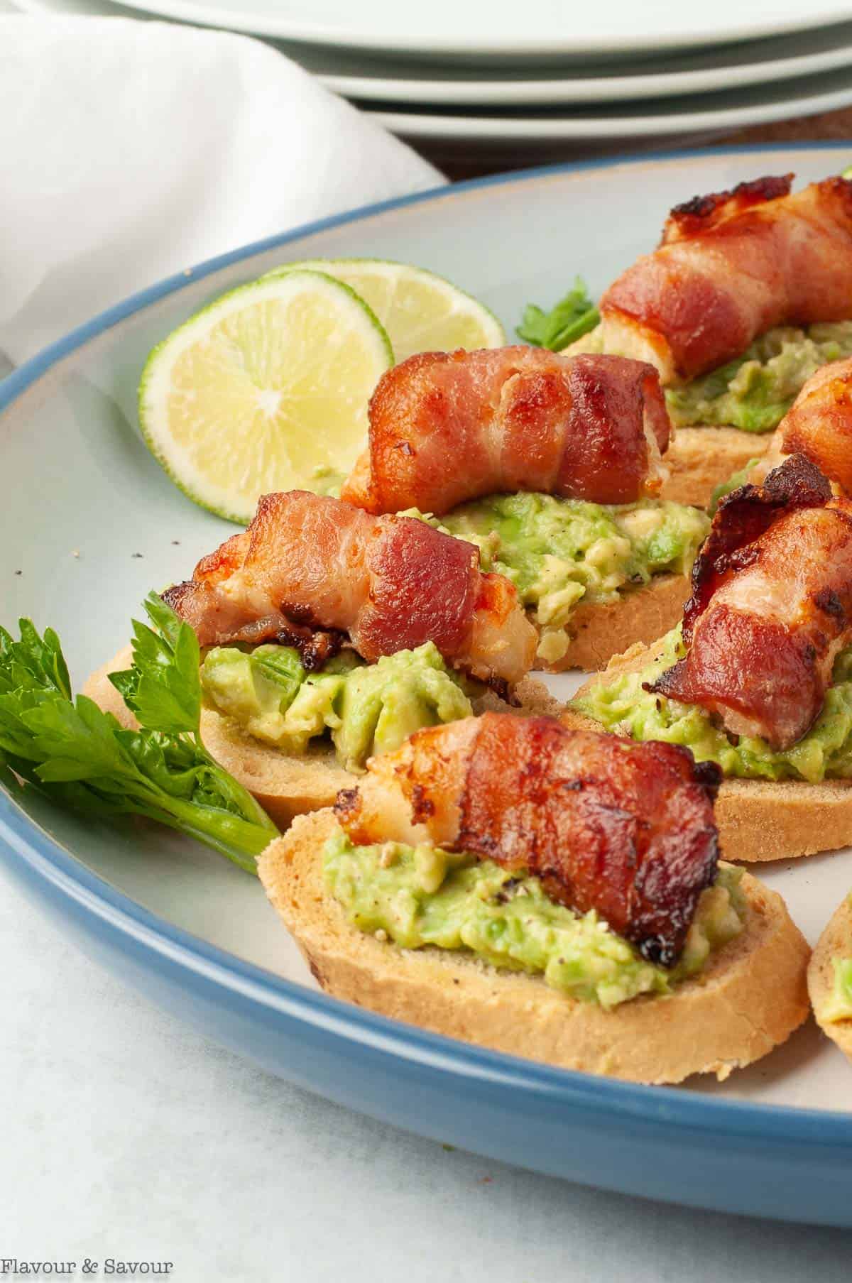 A plate filled with Bacon-Wrapped Shrimp (or Prawn) Tapas, a crostini topped with avocado and a bacon wrapped shrimp.