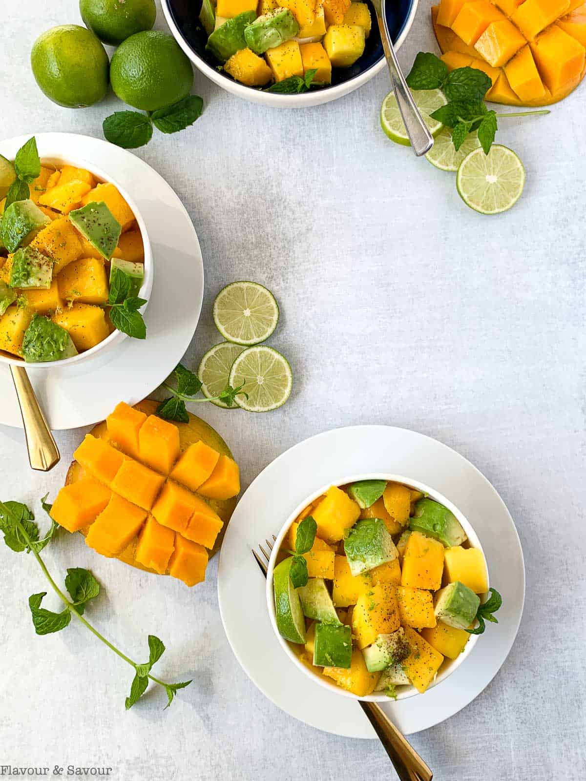 Overhead view of 3 bowls of Mango Avocado Chili Lime Salad with extra limes, lime slices and cubed mango on the surface.