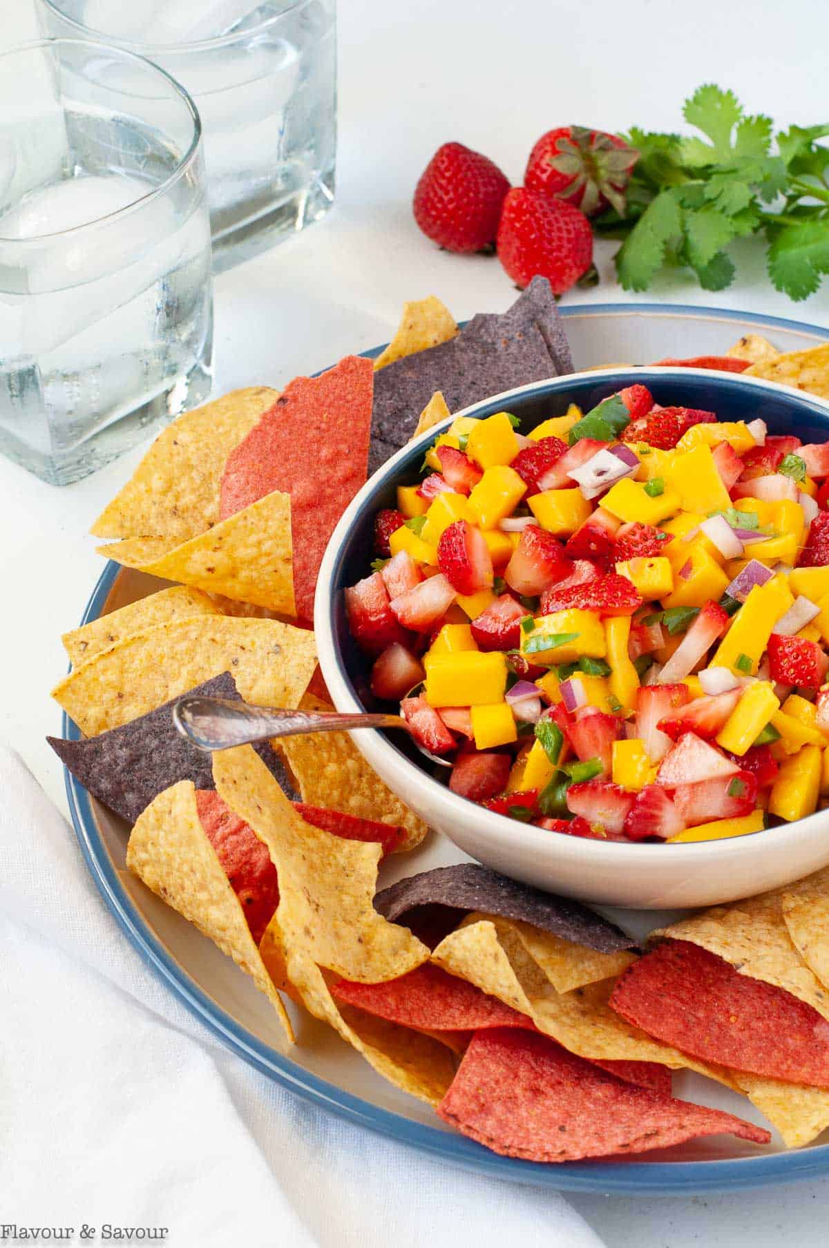 A bowl of strawberry mango salsa with tortilla chips and fresh strawberries and cilantro in the background.