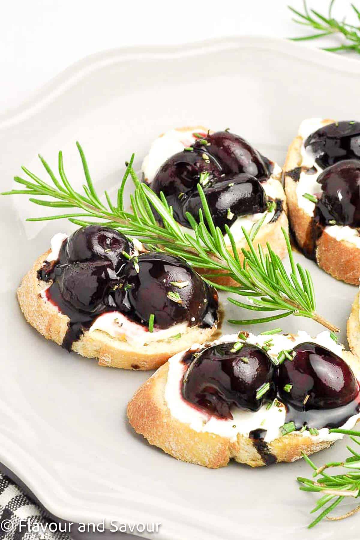 Roasted cherry crostini with goat cheese on a plate with a rosemary sprig.
