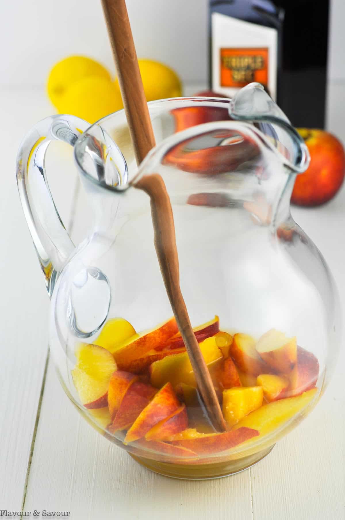 A pitcher with fresh sliced peaches, lemon juice and orange liqueur with a wooden spoon.