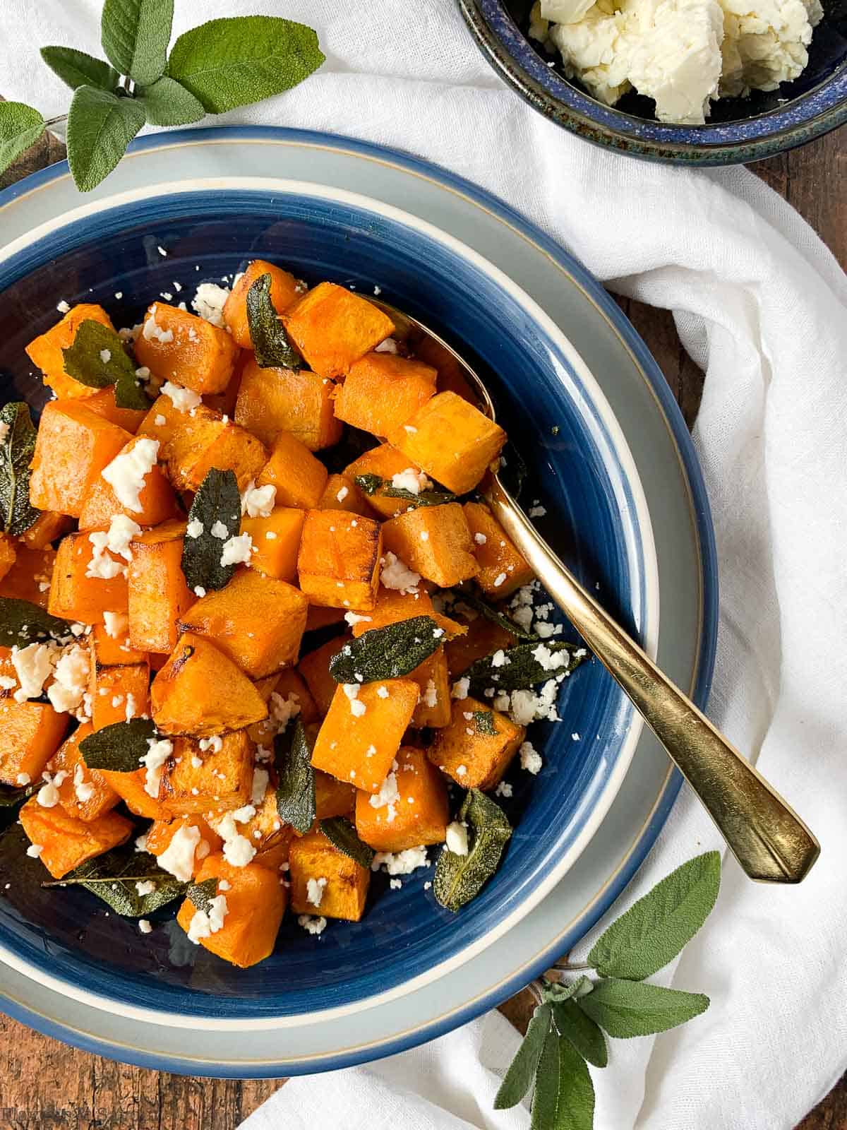 Overhead view of a bowl of butternut squash with feta cheese and toasted sage leaves