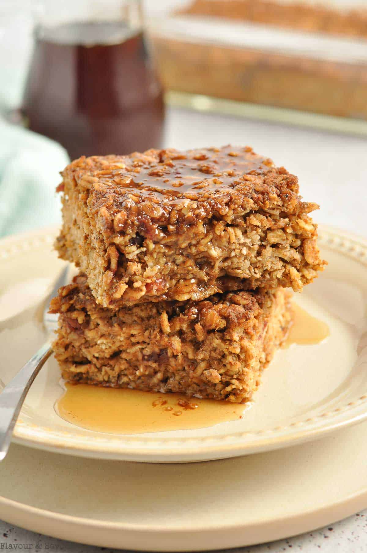 Two squares of cranberry apple pumpkin baked oatmeal with a pitcher of maple syrup.