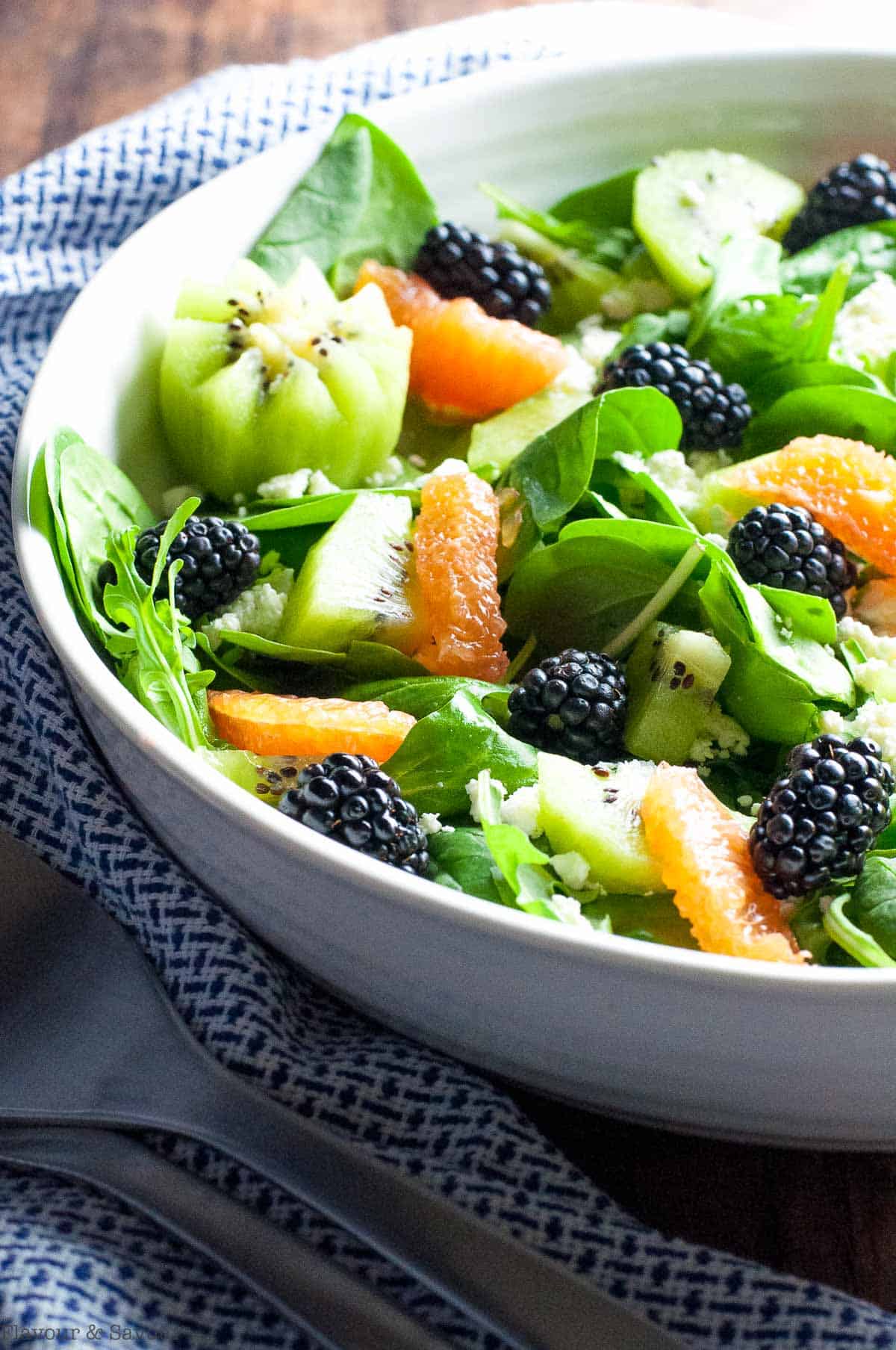 side view of blackberry kiwi salad with a pair of salad servers nearby.