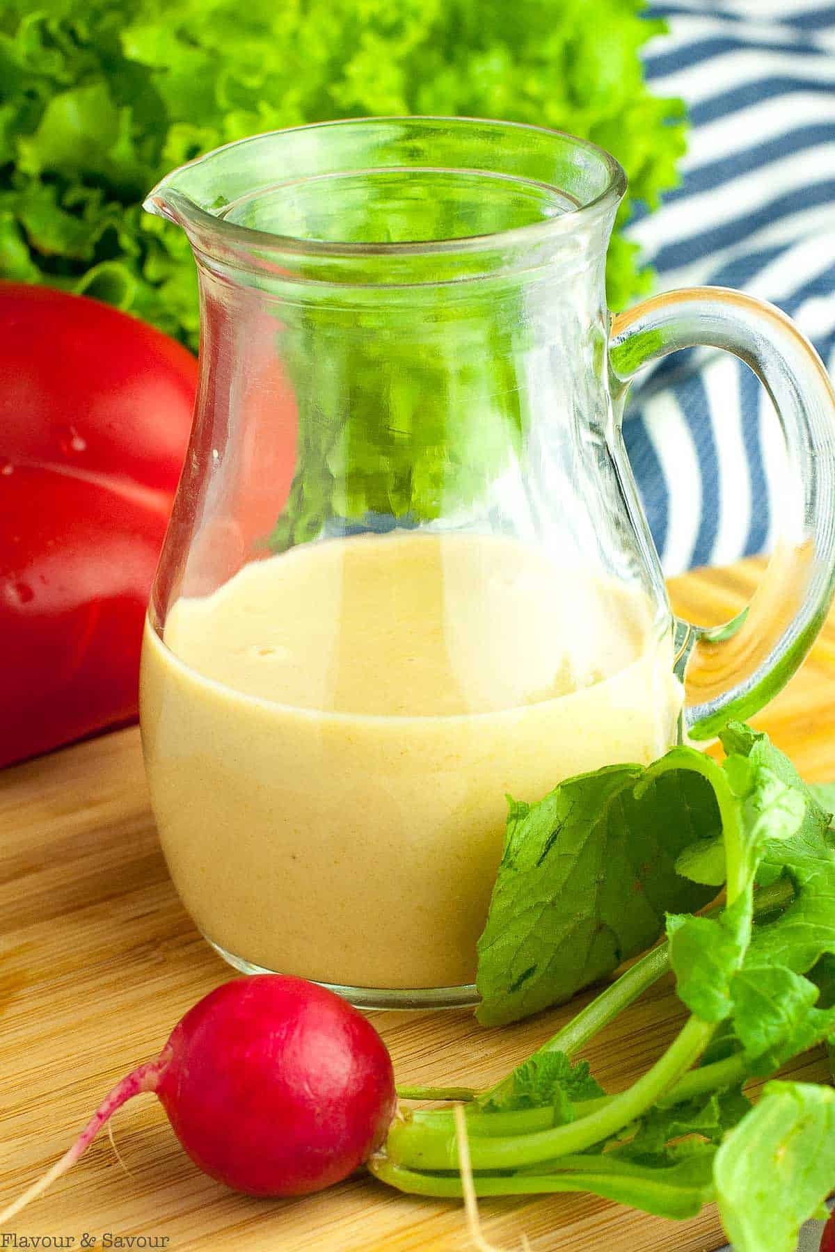 A small pitcher of lemon tahini dressing with fresh salad ingredients beside.