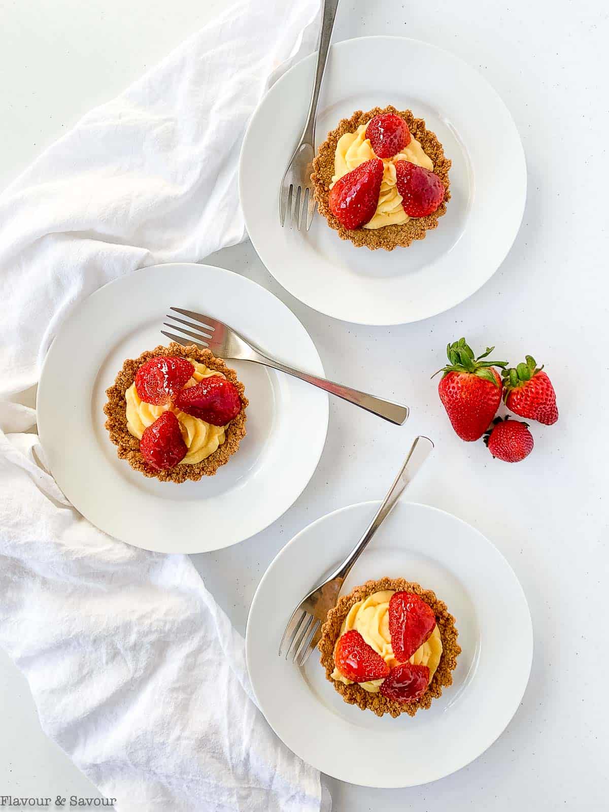 Three strawberry tarts with graham cracker crust and pastry cream on white plates.