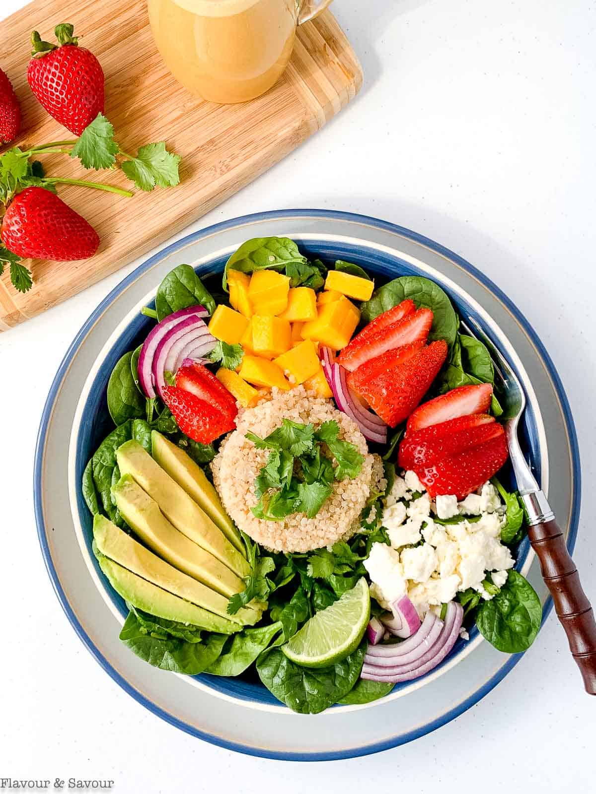 A bowl of strawberry mango quinoa salad with a pitcher of dressing and fresh strawberries.