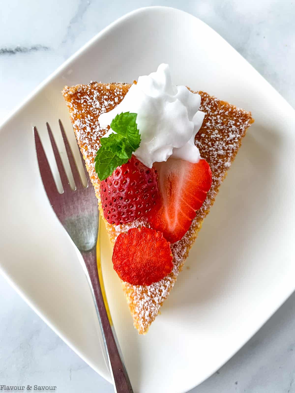 Overhead view of a slice of flourless lemon almond cake topped with strawberries and whipped cream.