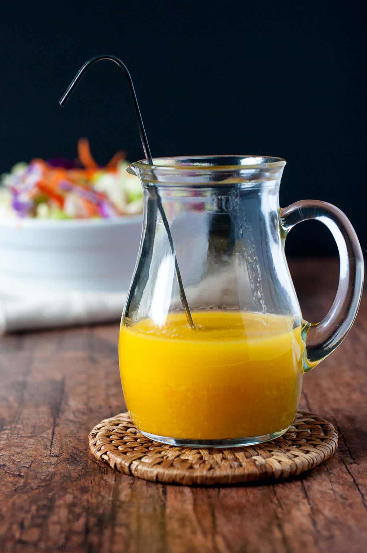 a small pitcher of ginger-orange dressing with a bowl of coleslaw in the background.