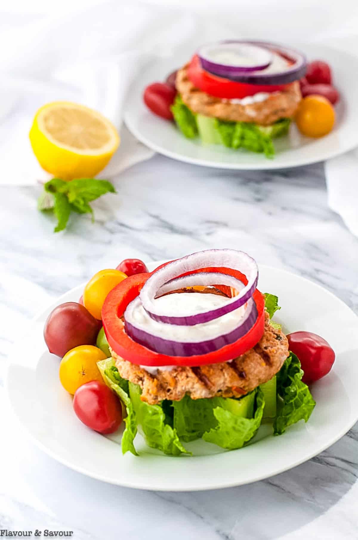 Two plates with open face harissa chicken burgers made with ground chicken and garnished with tomato, lettuce and onion.