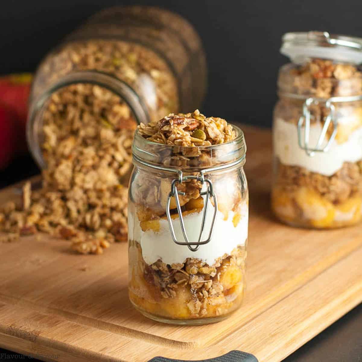 Two jars of apple crumble breakfast parfait with a large jar of granola in the background.