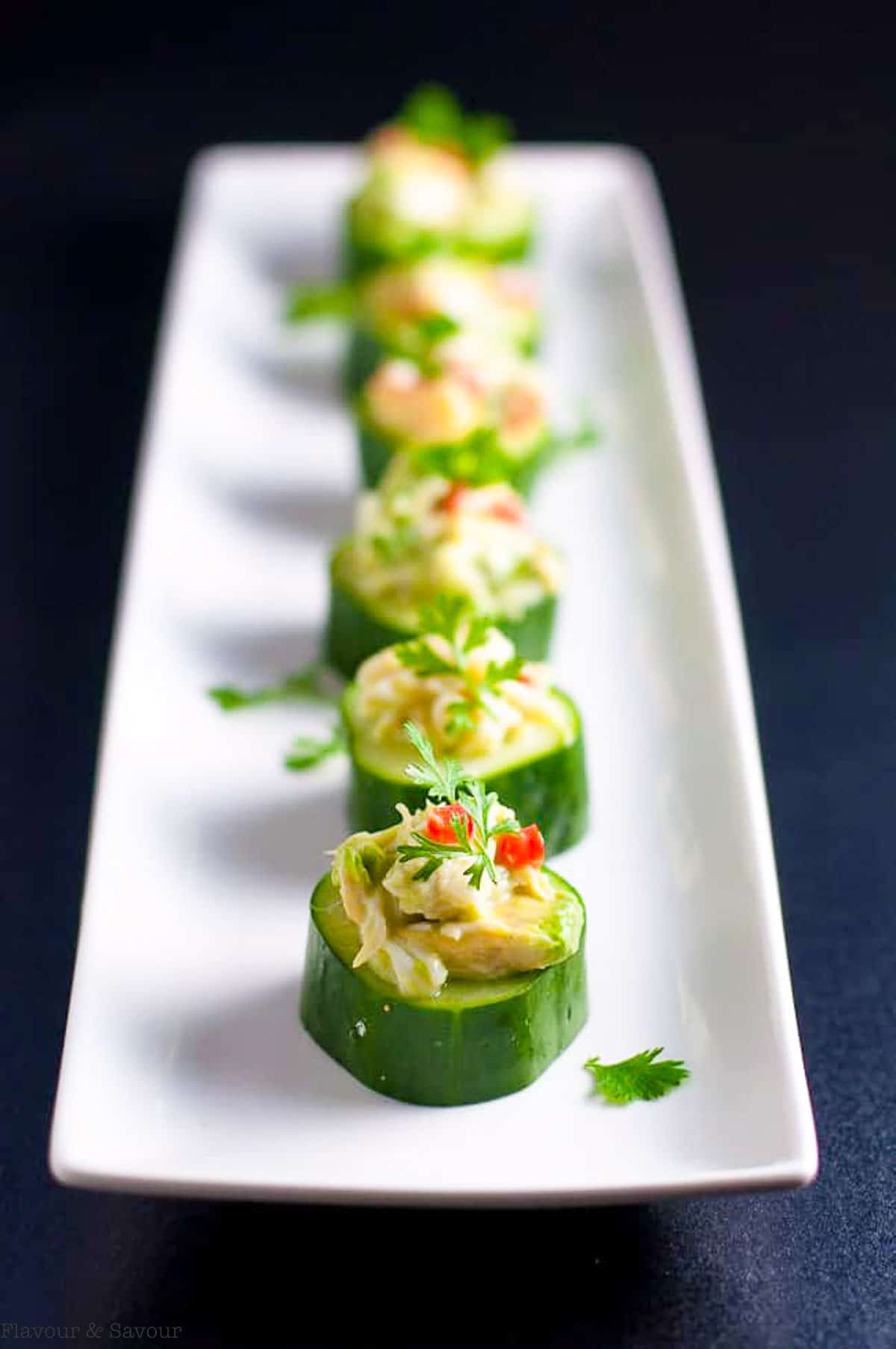 Crab stuffed cucumber bites in a single row on a narrow white platter.
