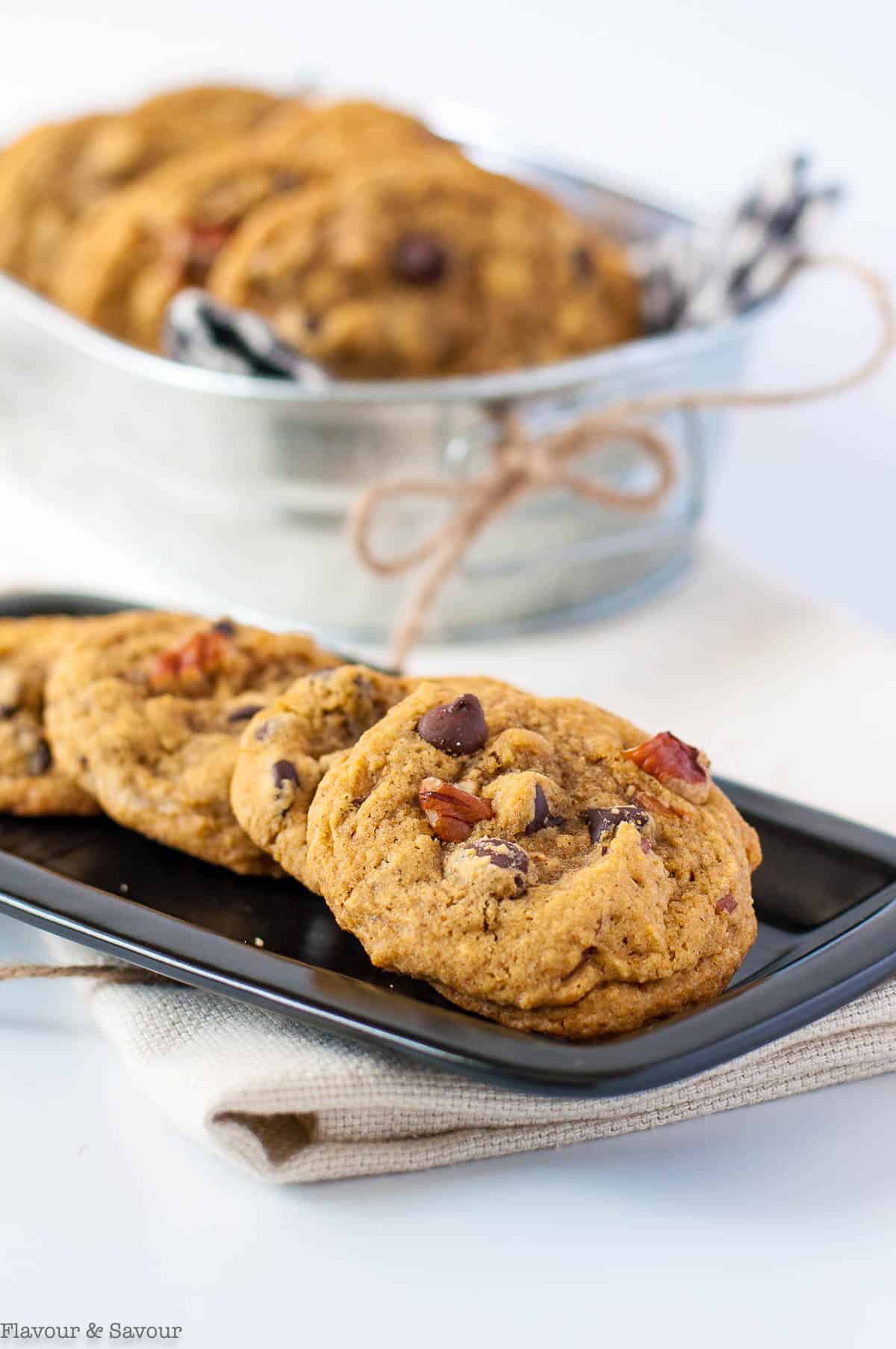 Pumpkin pecan chocolate chip cookies on a rectangular black plate with a metal basket of cookies in the background.