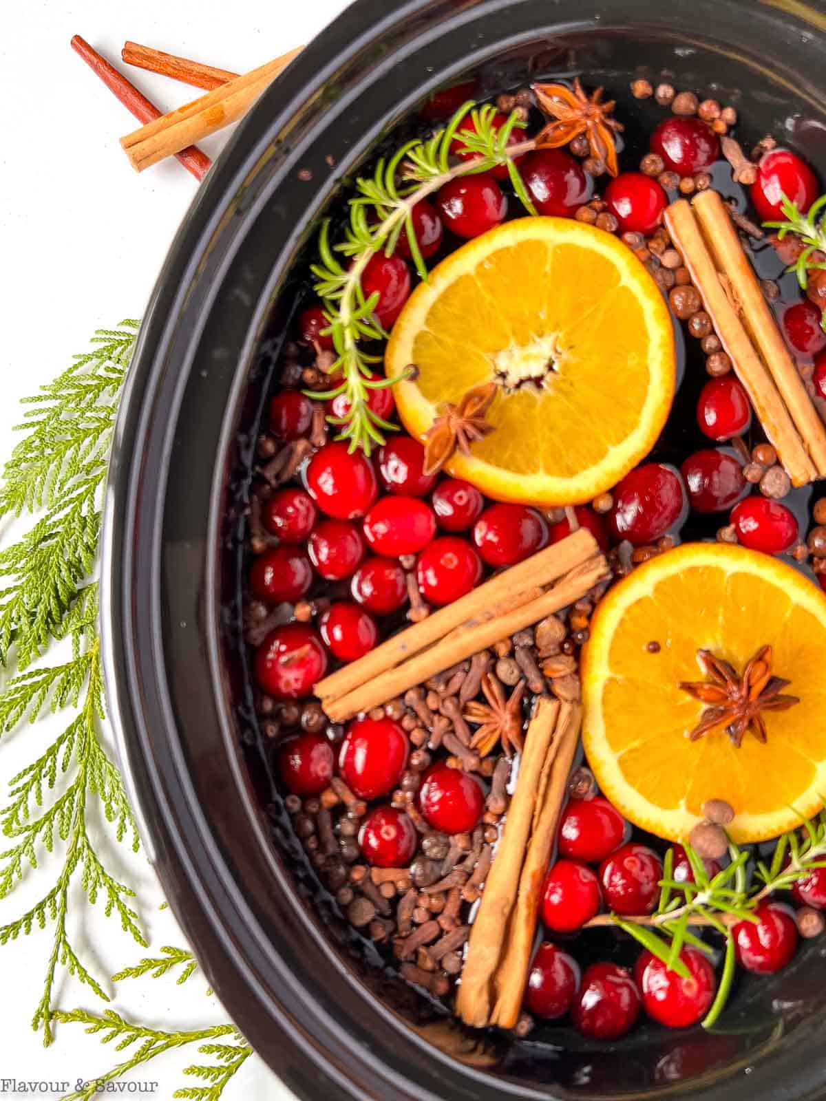 Close up overhead view of spices, cranberries and oranges in a slow cooker bowl.