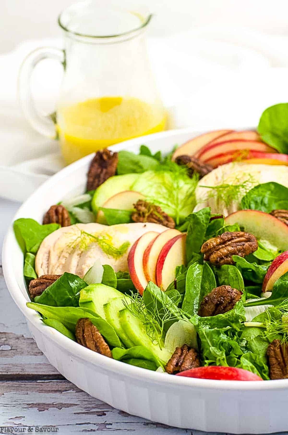 A small pitcher of lemon vinaigrette beside a shallow bowl filled with fennel apple chicken salad.