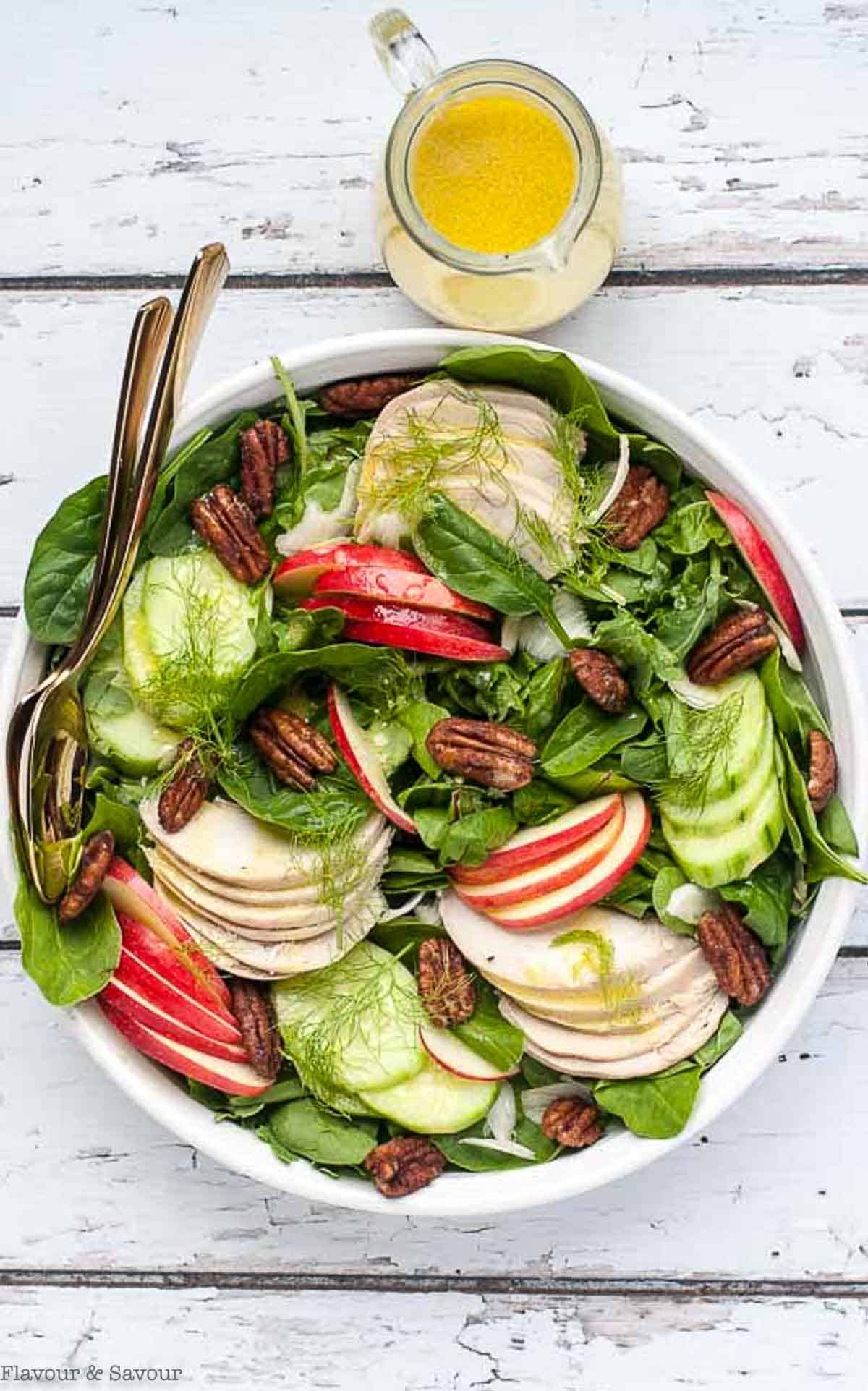 Overhead view of a large shallow serving bow with a salad made with spinach, apples, chicken, fennel and pumpkin spice pecans.