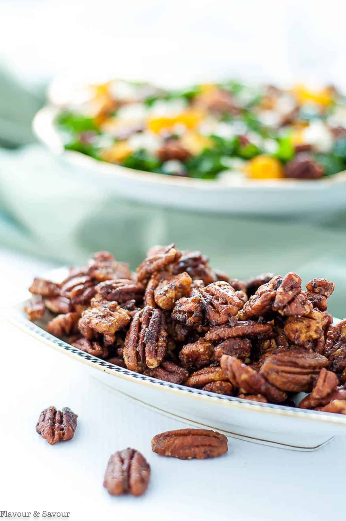 A serving dish of caramel spiced pecans with a salad in the background.
