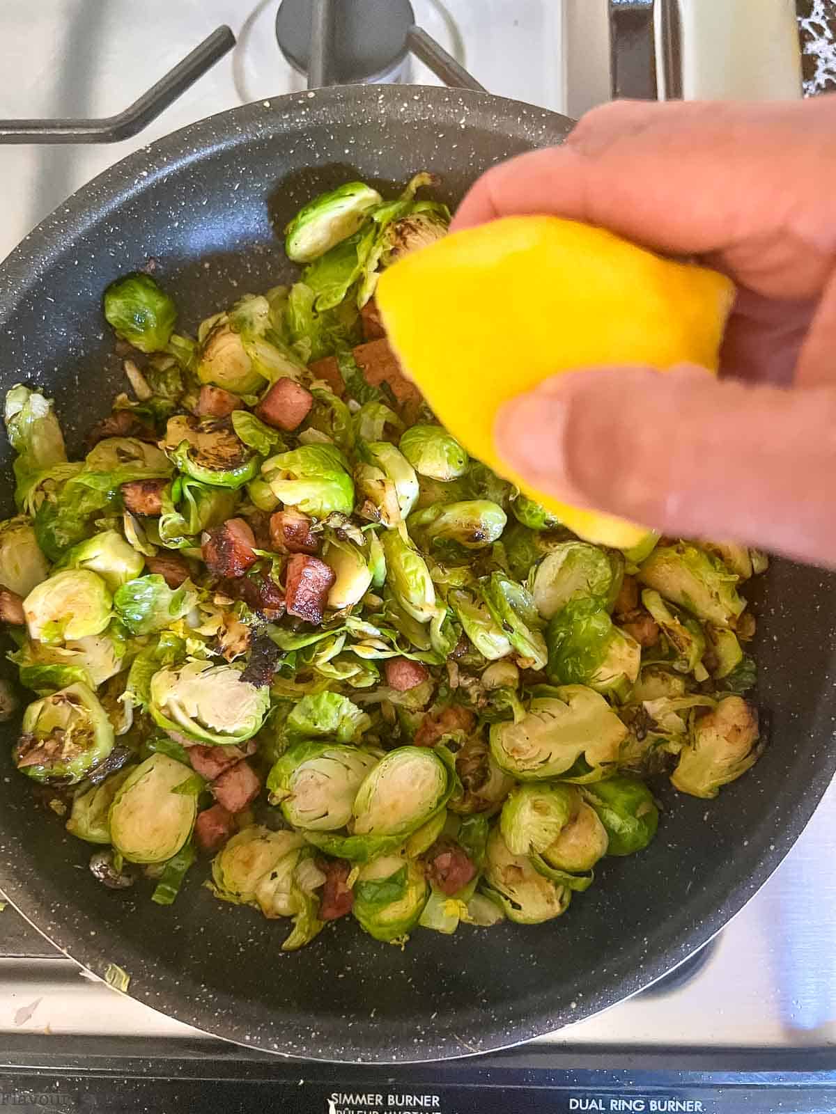 Squeezing fresh lemon juice on sautéed Brussels sprouts and pancetta in a skillet.