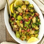 Overhead view of a bowl of sauteed Brussels sprouts with pancetta garnished with lemon wedges.