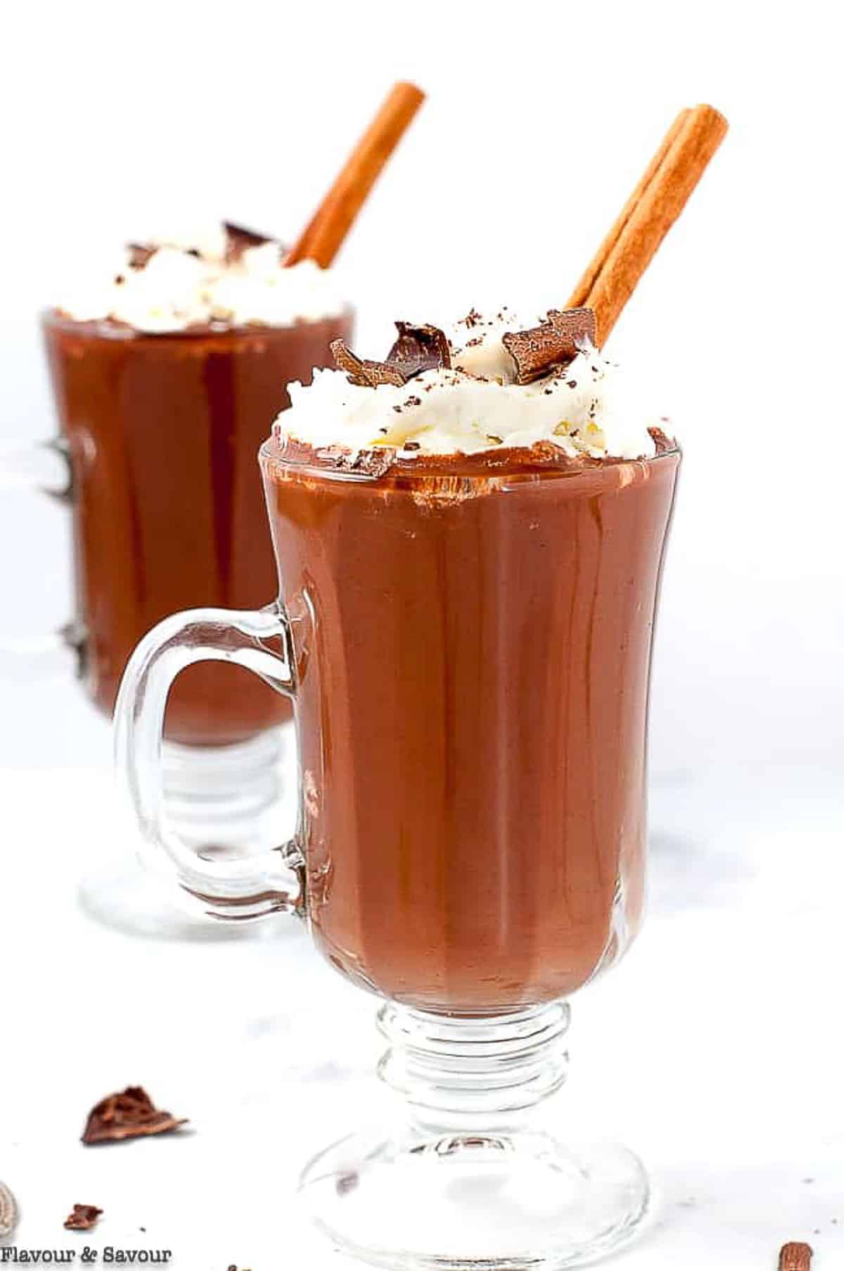 Two glass mugs of Dairy-Free Mexican Hot Chocolate topped with whipped cream, chocolate shavings and cinnamon sticks.