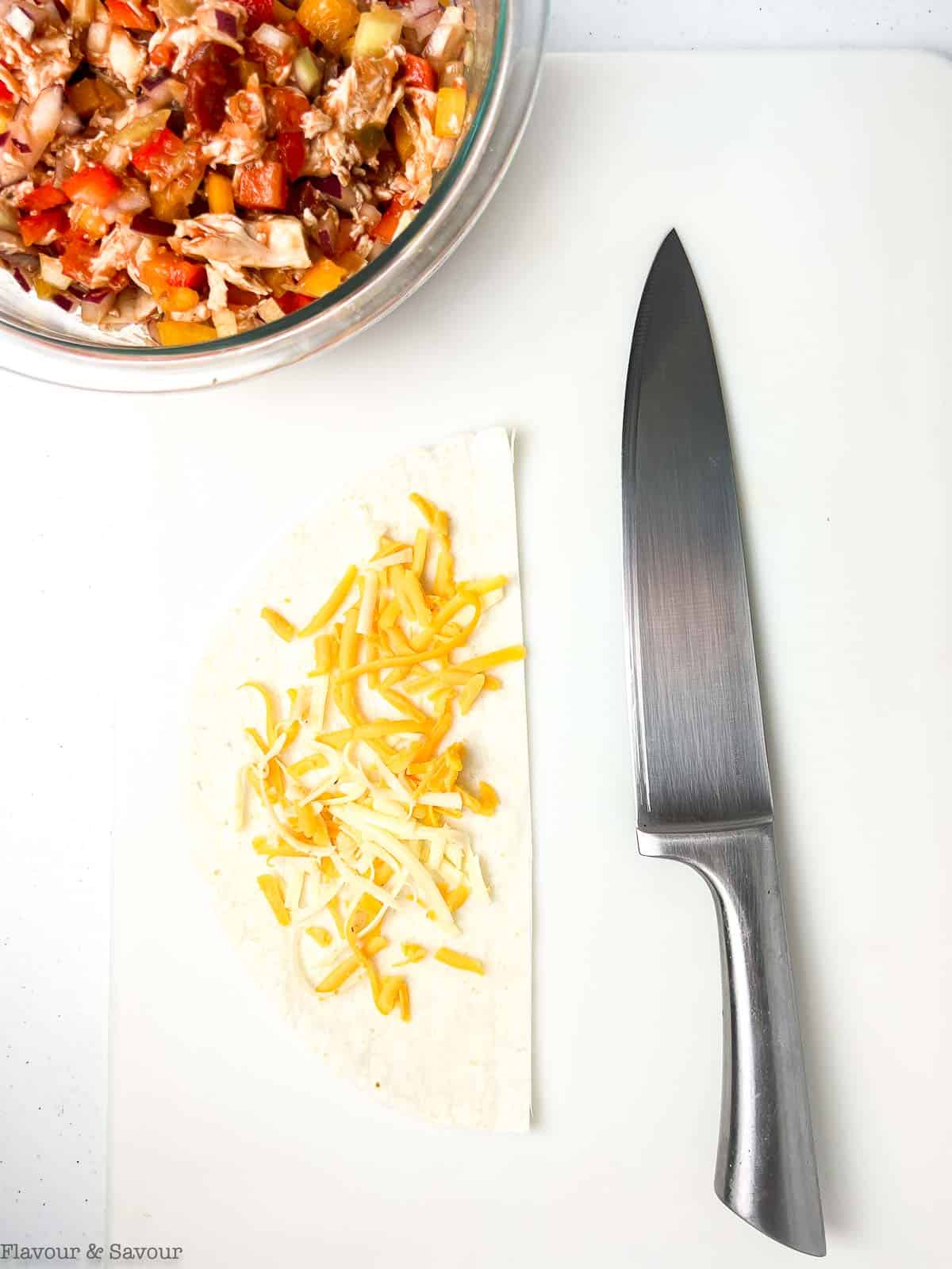 Half a tortilla on a cutting board with a knife and a bowl of chicken mixture.