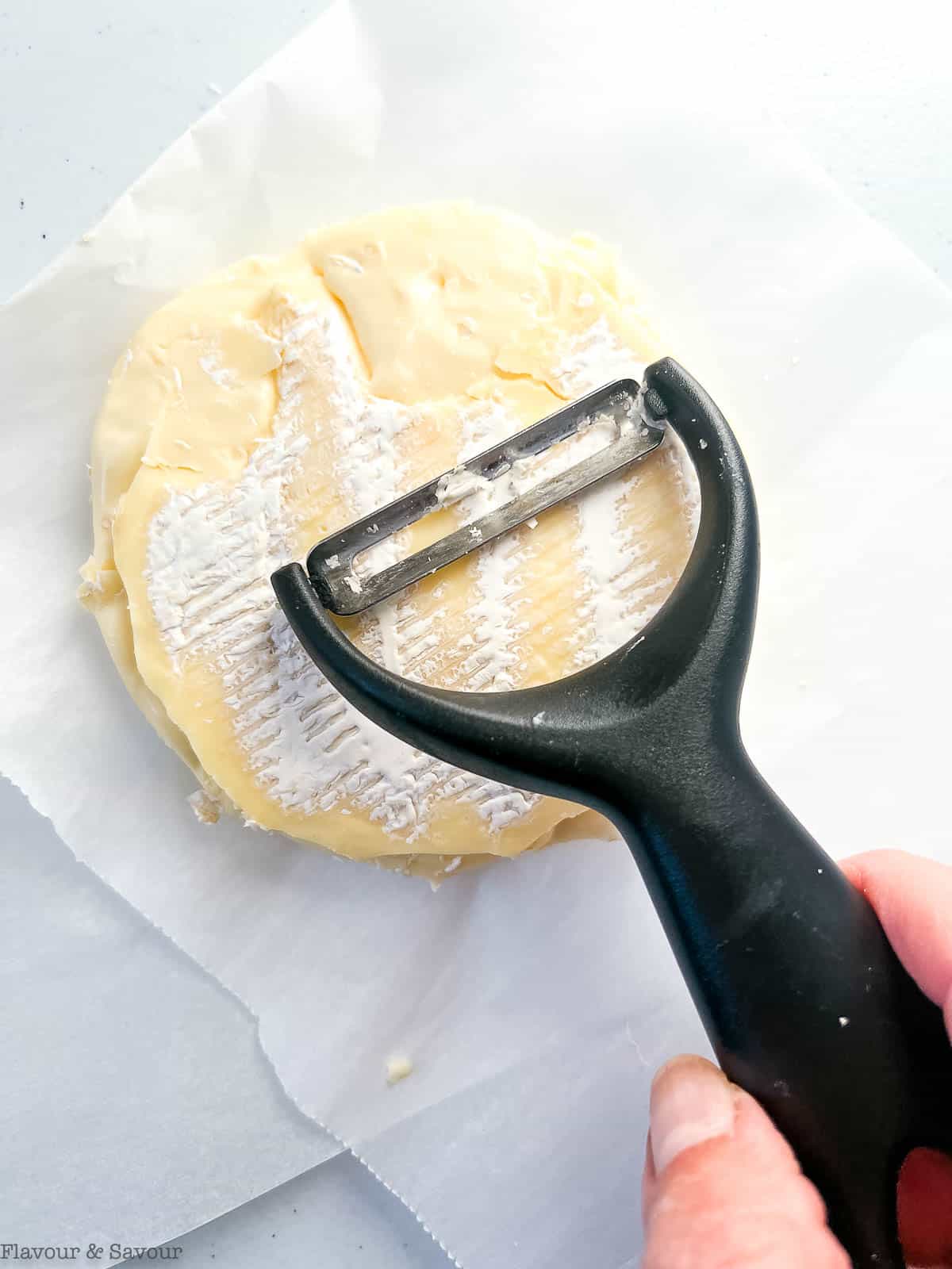 Removing rind from a wheel of Brie with a vegetable peeler.