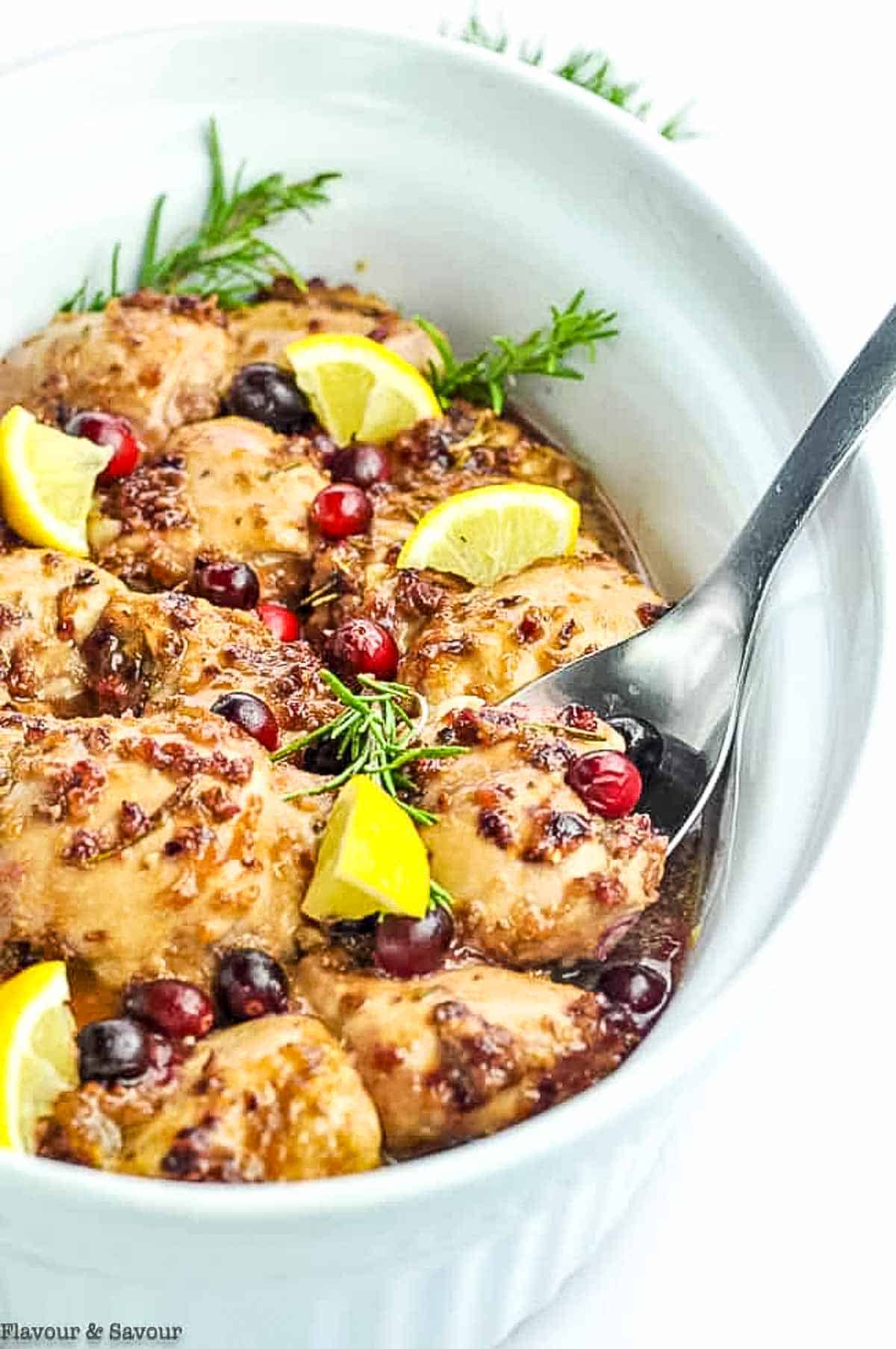 A white serving bowl with cranberry baked chicken thighs with rosemary and lemon.