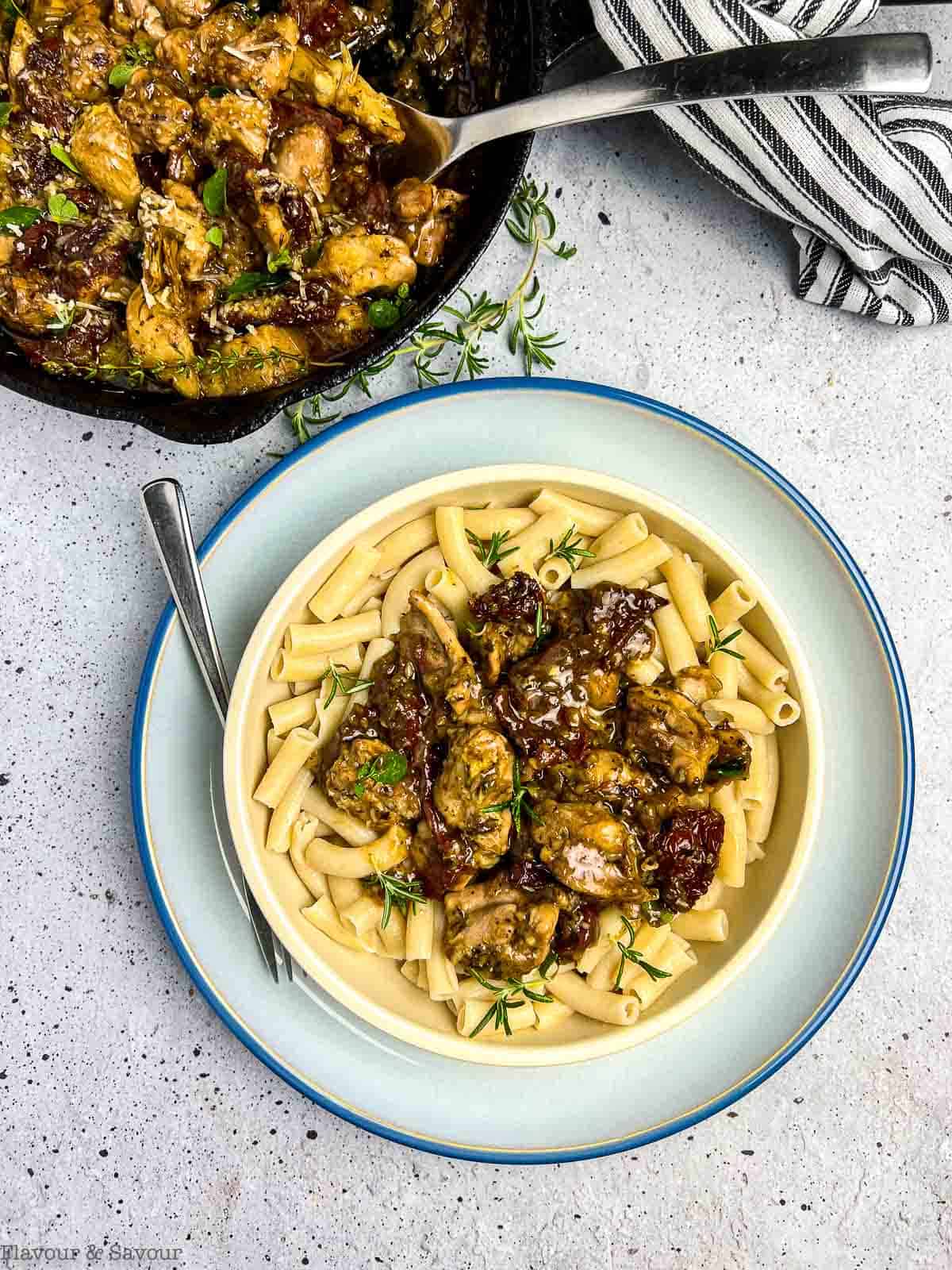 A bowl of sun-dried tomato artichoke chicken on gluten-free pasta beside a cast iron skillet.