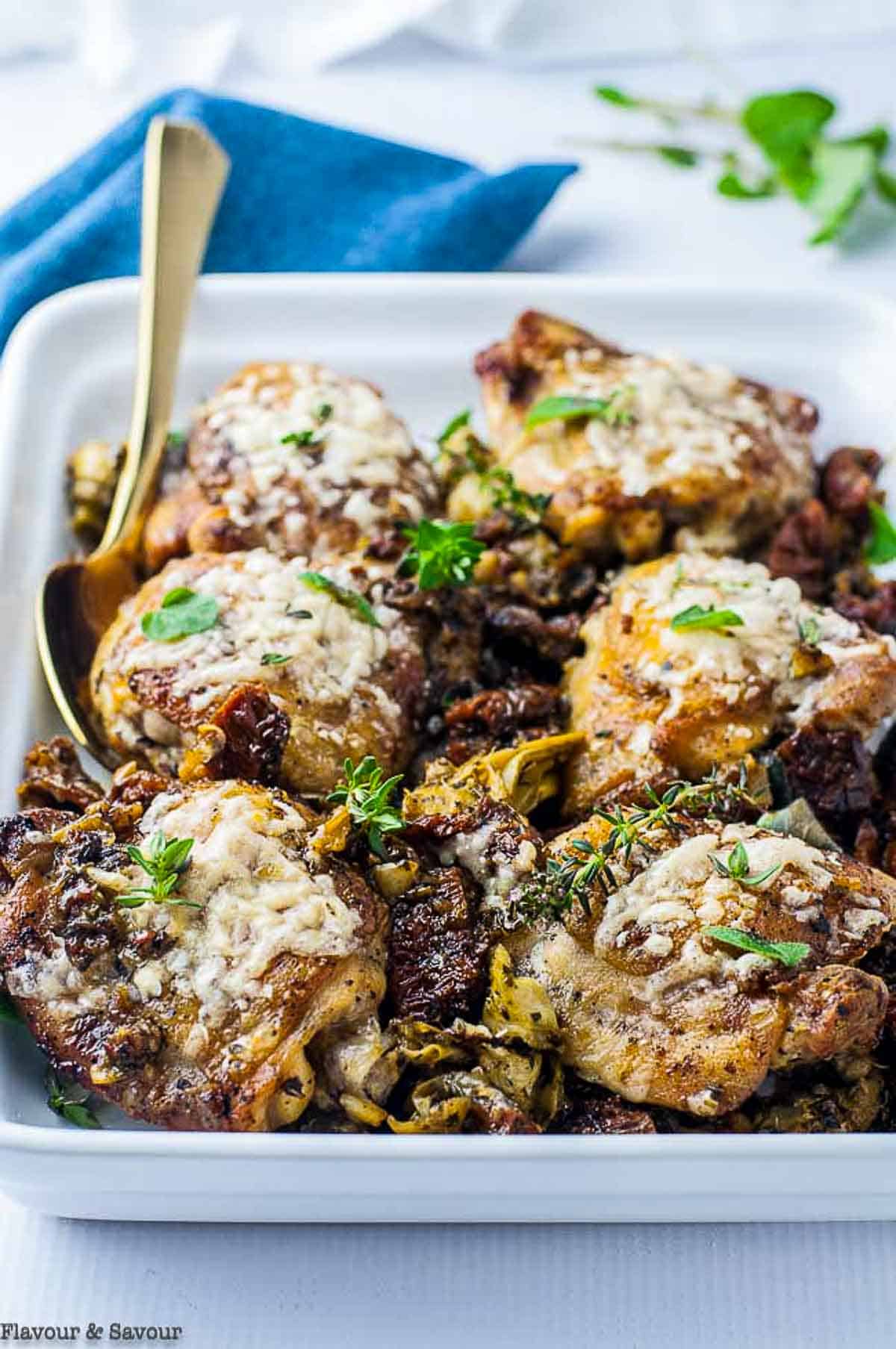 Bone-in skin-on chicken thighs with sun-dried tomatoes and artichokes in a white serving dish.