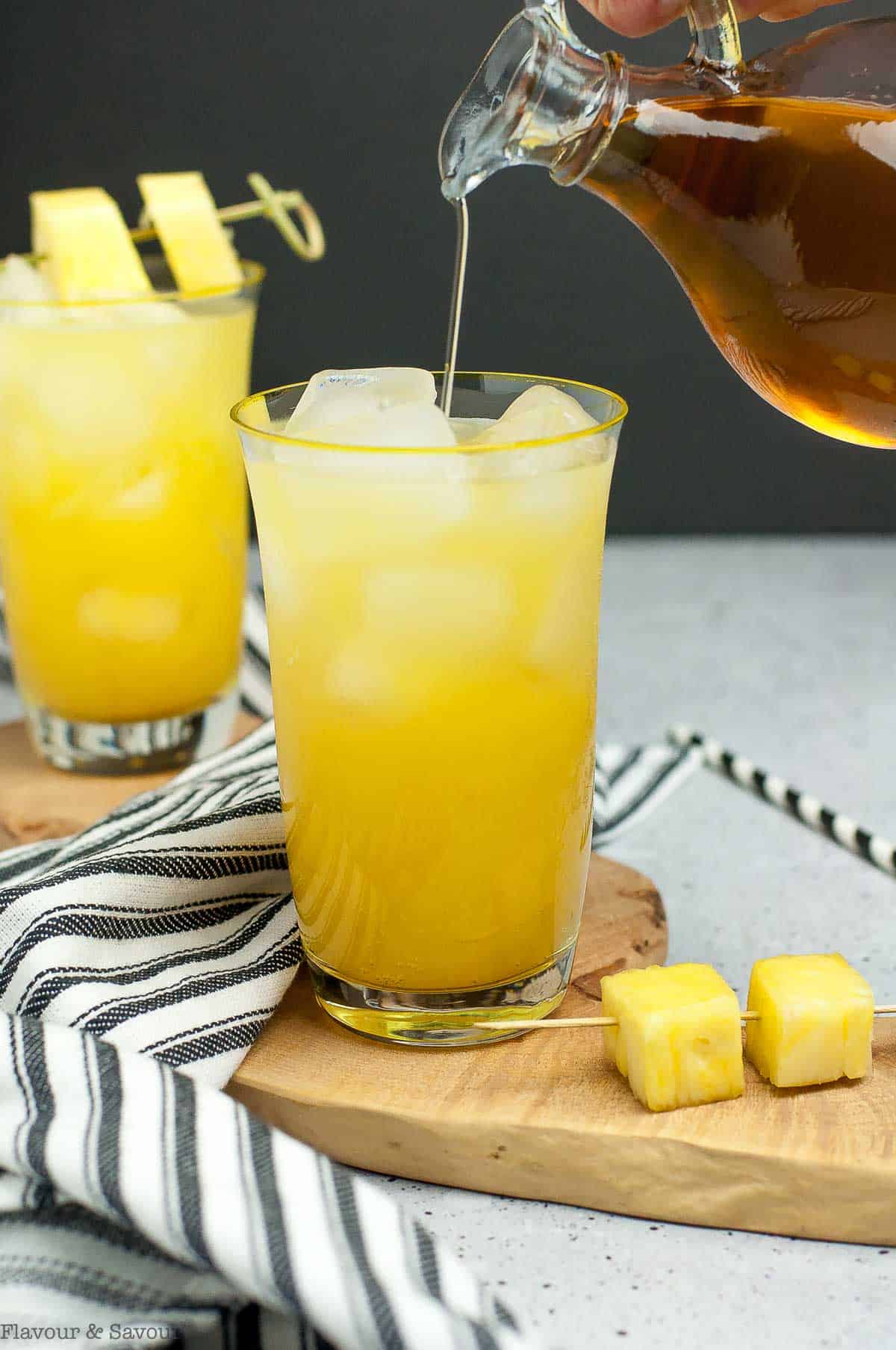 Pouring a tablespoon of cinnamon simple syrup into a glass of pineapple ginger mocktail.