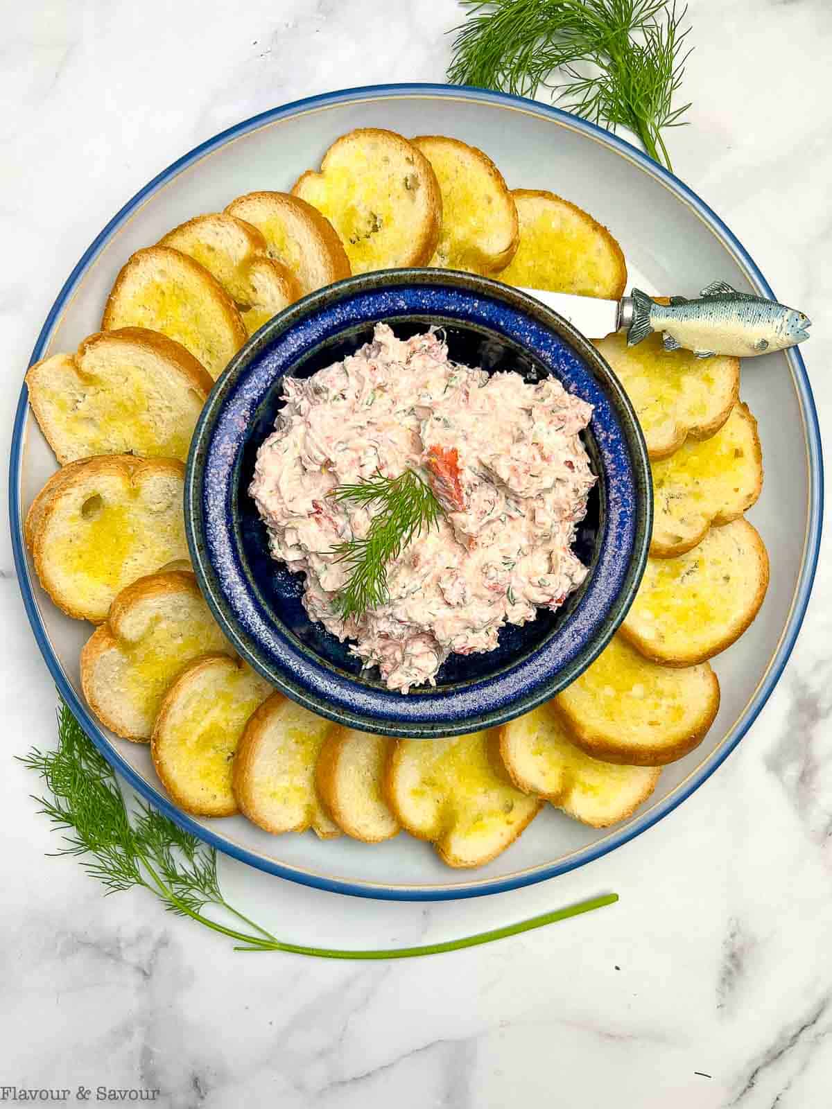 Overhead view of a bowl of smoked salmon dip with crostini on a plate around the bowl.