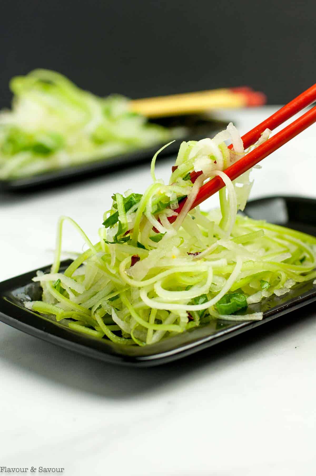 A pair of red chopsticks lifting Asian Pear Slaw from a small black plate.
