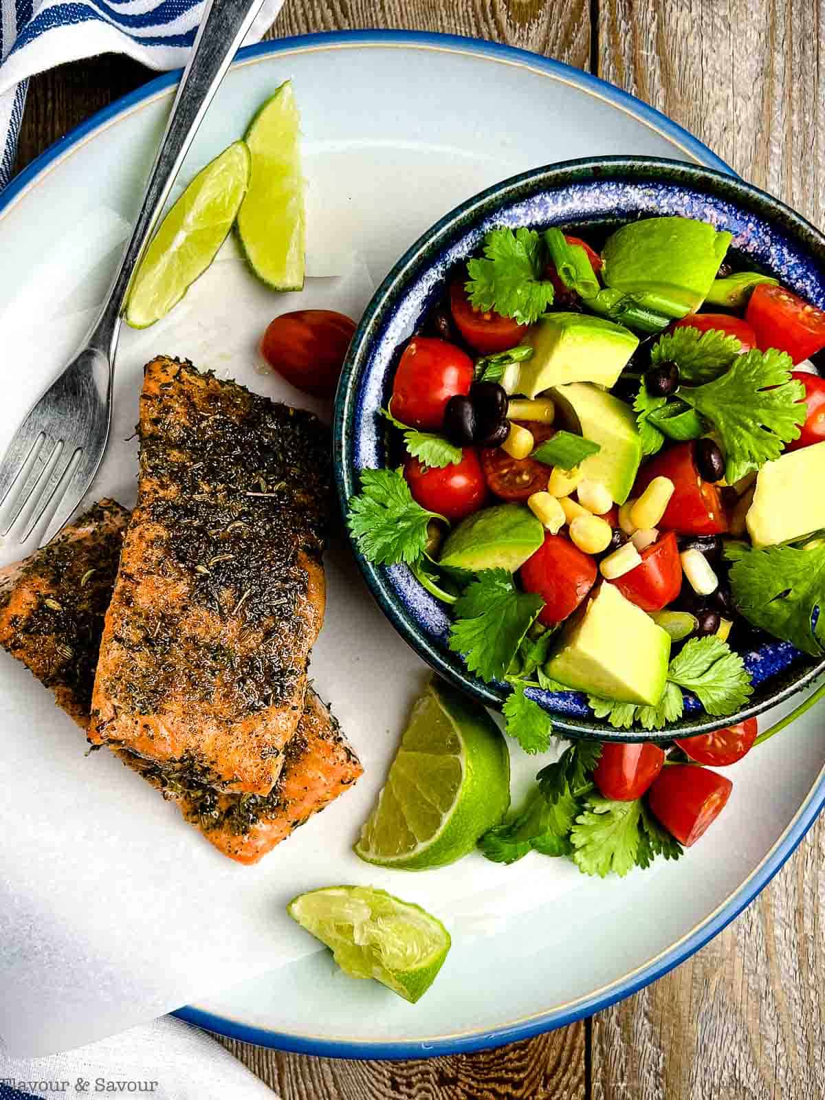 Overhead view of a plate with two fillets of jerk salmon and a bowl of corn and black bean salad.