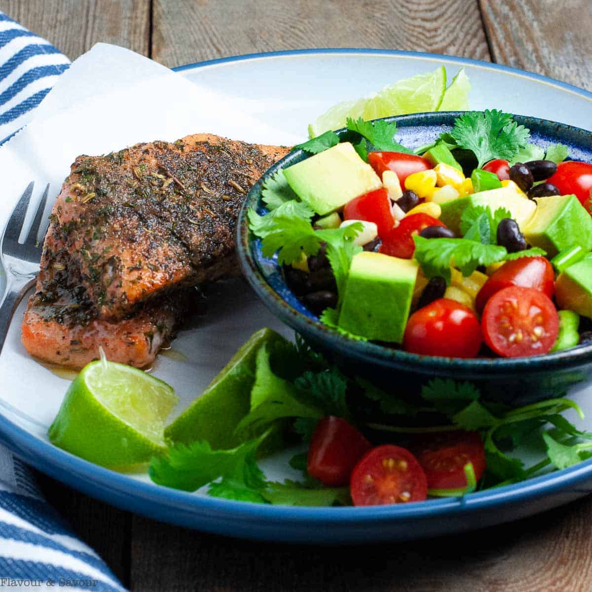 Jerk salmon and corn and black bean salad on a plate with lime wedges and cilantro leaves.