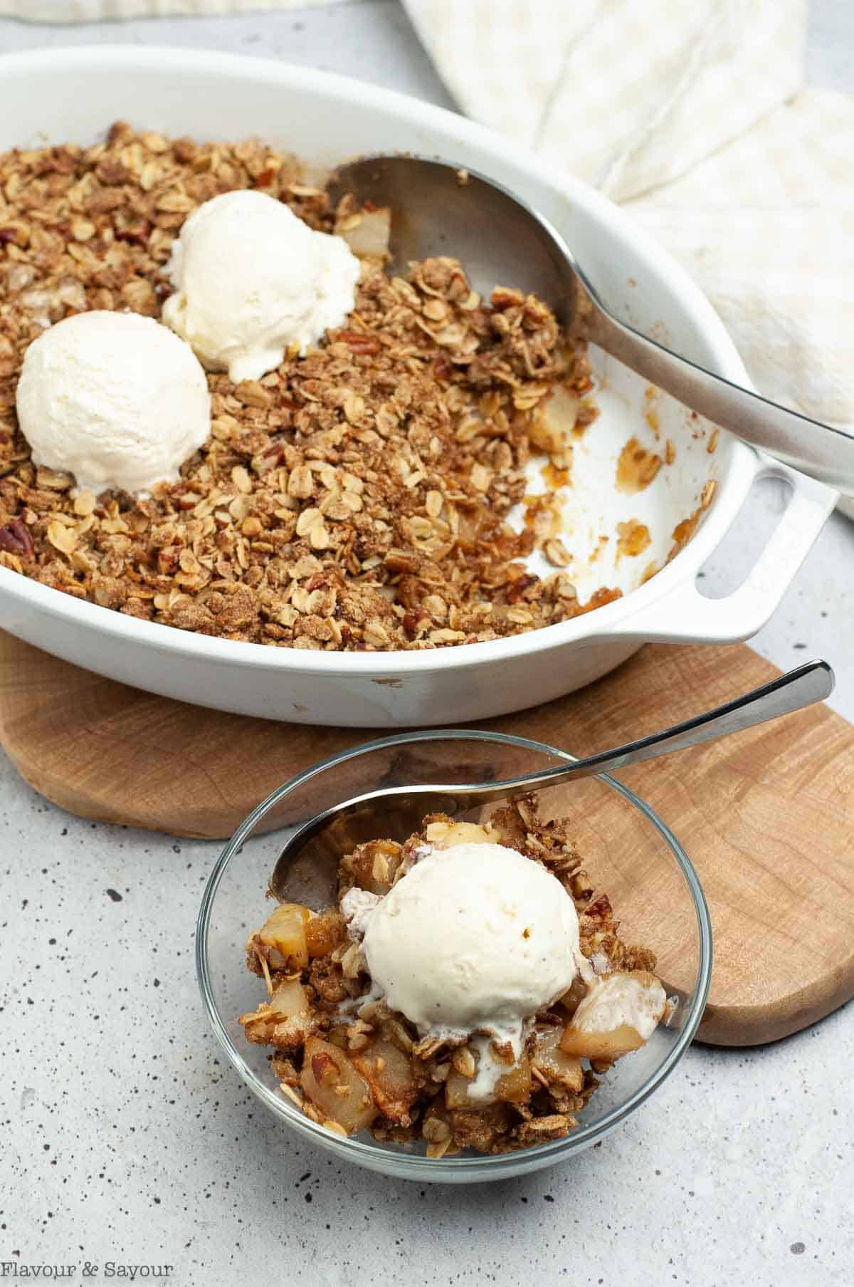 Overhead view of a bowl of pecan pear crisp with ice cream with a baking dish in the background.