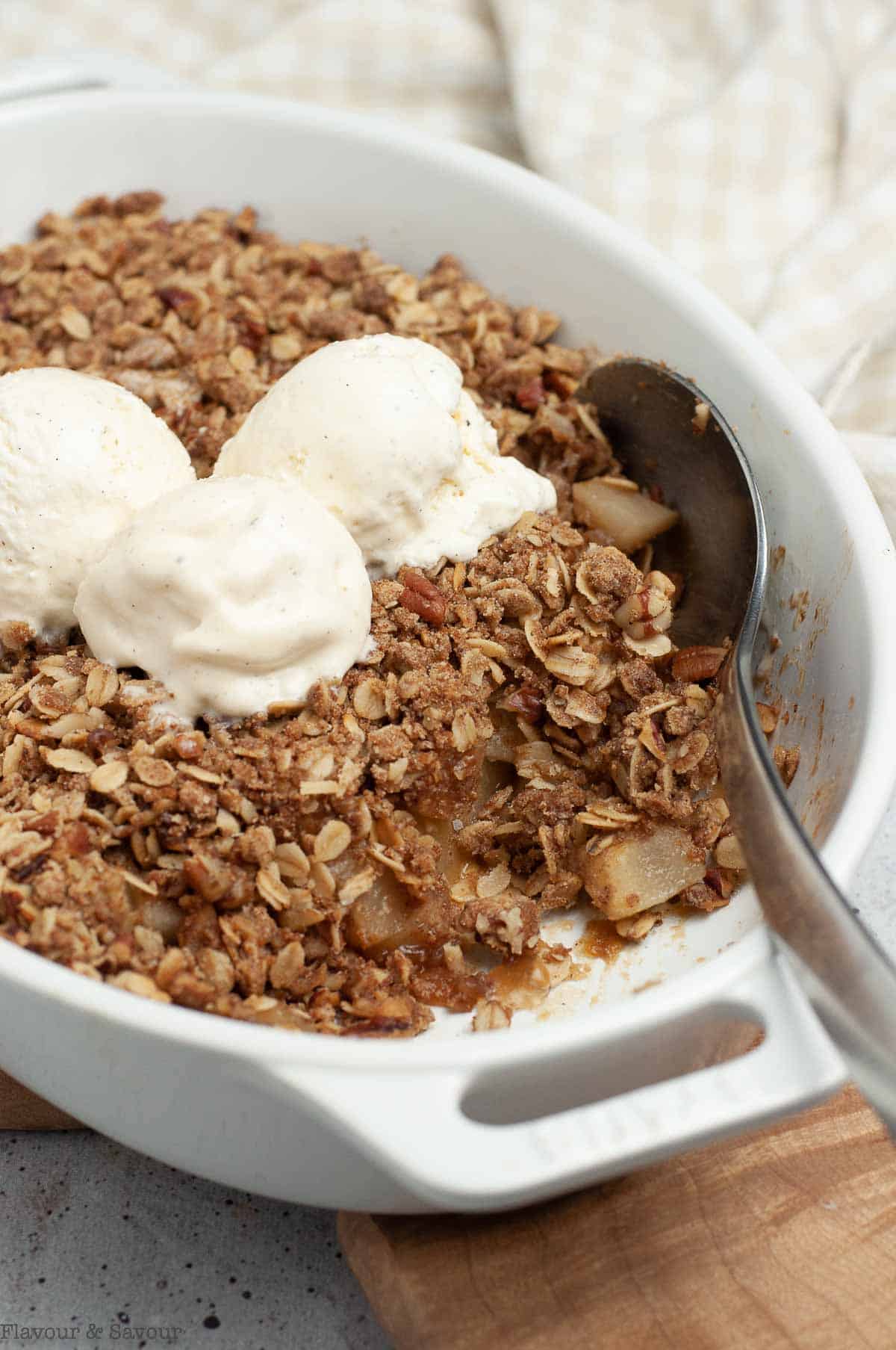A white oval baking dish with pecan pear crisp with 3 scoops of ice cream on top.