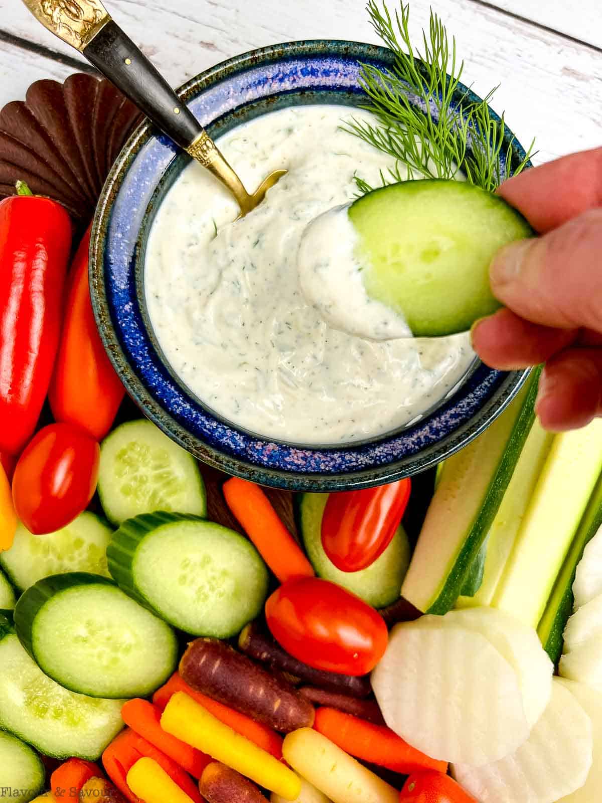 A platter of fresh raw vegetables with a bowl of Greek Yogurt Dill Dip and a hand dipping a slice of cucumber.