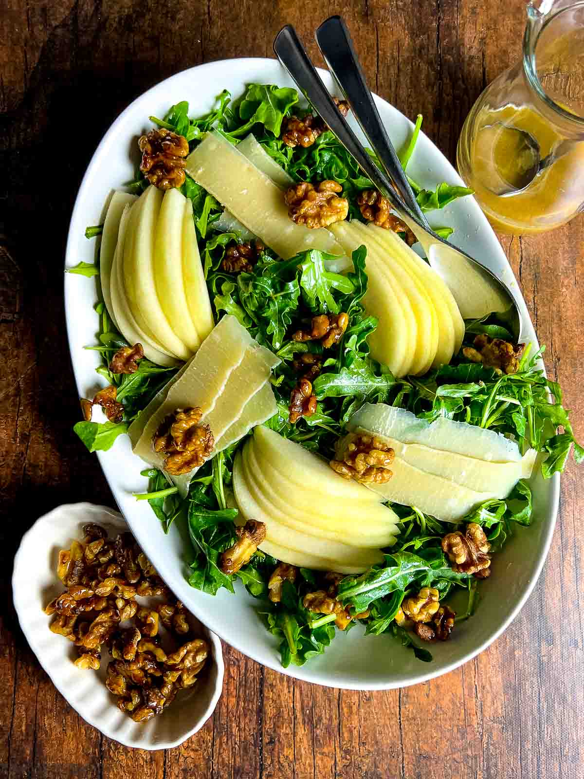 An oval bowl with arugula salad topped with sliced pear, shaved Parmesan and maple-glazed walnuts.