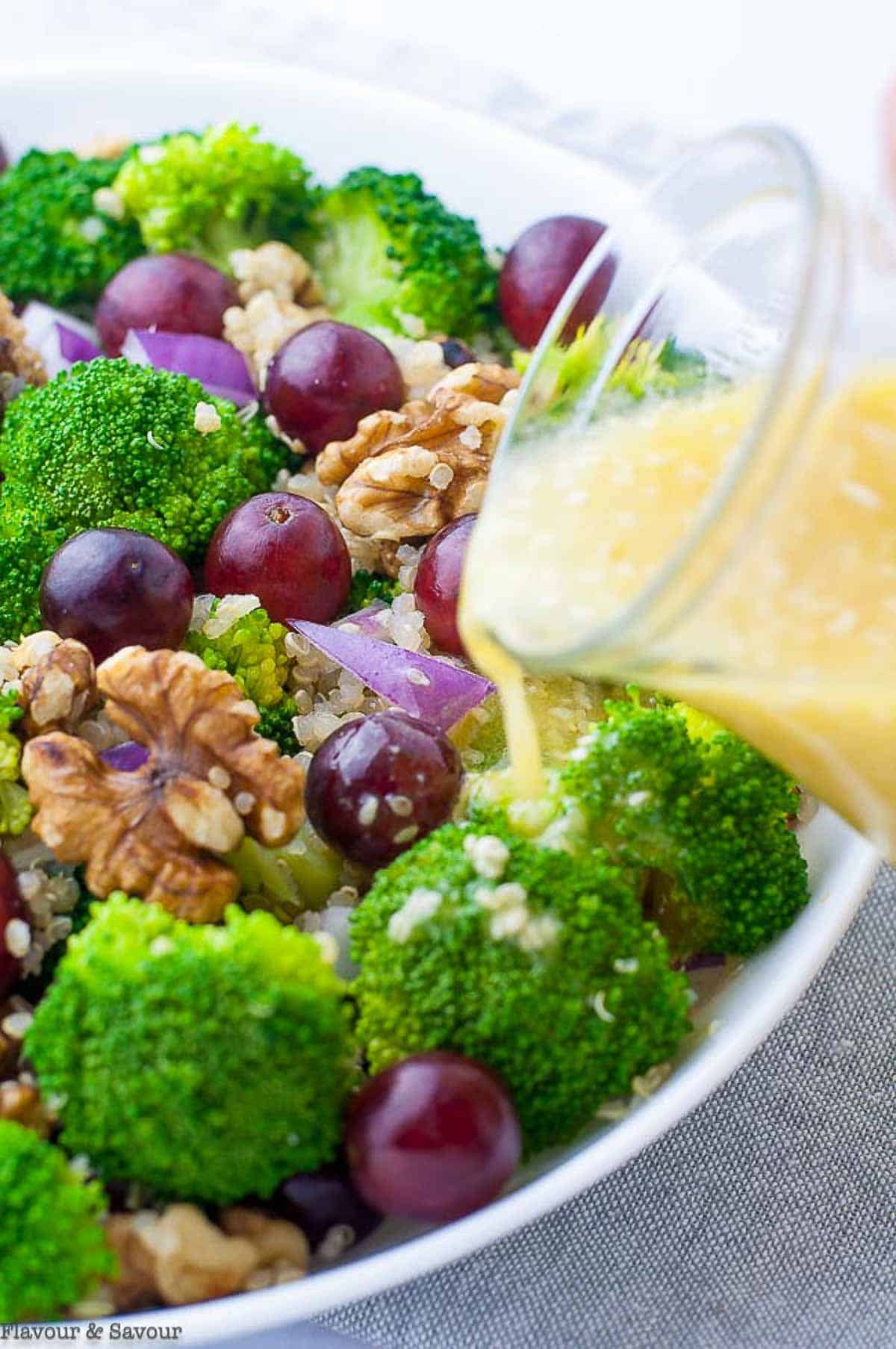Pouring dressing on a bowl of broccoli quinoa salad with red grapes.