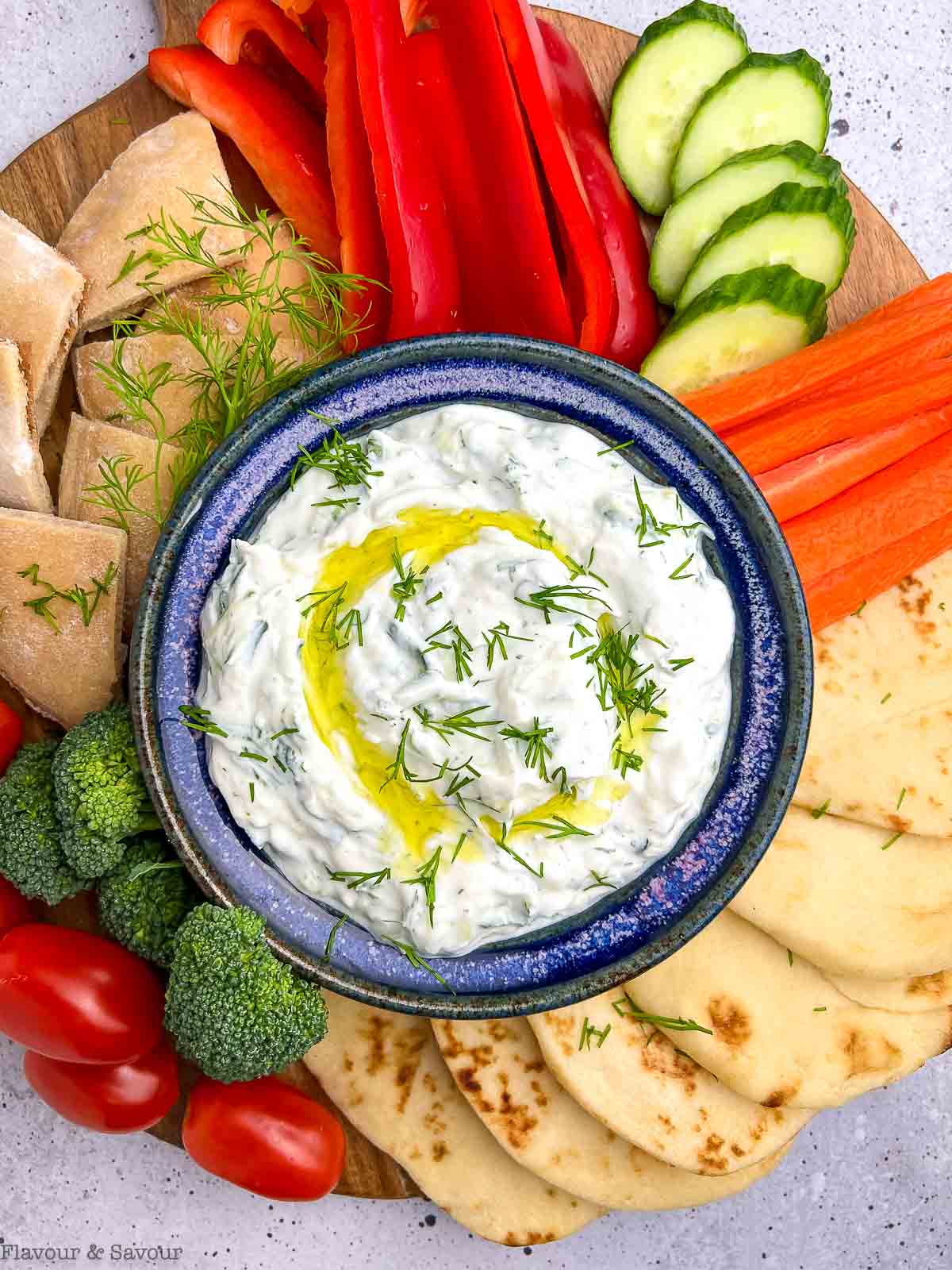 Overhead view of a bowl of tzatziki sauce surrounded by raw vegetables and pita bread.