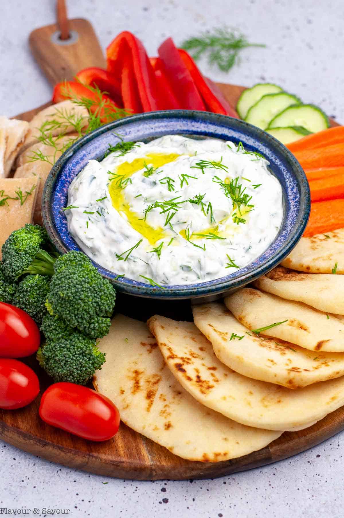 Greek Tzatziki sauce in a pottery bowl surrounded by raw vegetables and pita bread.